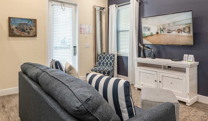 Two Bedroom Villa Cozy seating area in a living room featuring a grey sofa, white TV console, wall-mounted television, and a patio door with blinds at Margaritaville Resort Orlando by Kenwood Travel