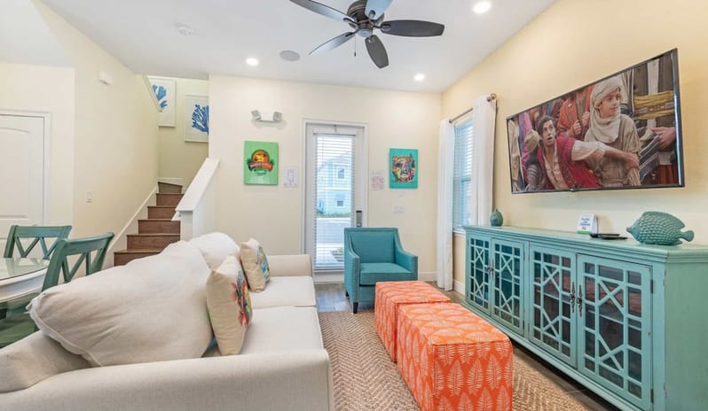 Two Bedroom Villa Spacious living area with white sofa, turquoise media console, large TV, and orange ottomans near a staircase at Margaritaville Resort Orlando by Kenwood Travel