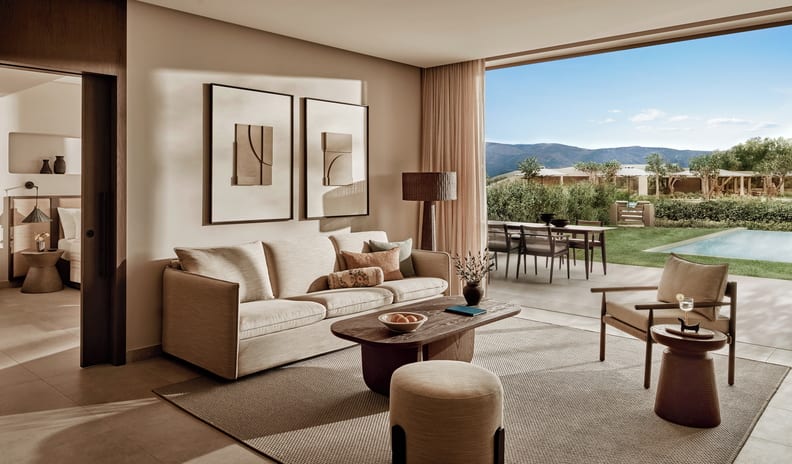 Deluxe Three Bedroom Suite Private Garden Pool View Minimalist modern living room featuring a cream sofa, dark wood coffee table, round ottoman, framed wall art, and open sliding glass door at Ikos Kissamos by Kenwood Travel