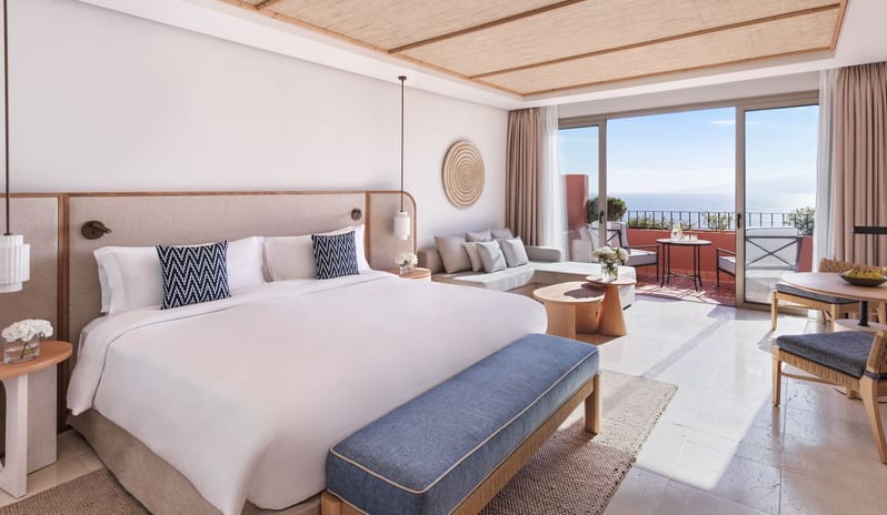 Citadel Two Bedroom Family Resort View Modern luxury resort suite with a king-size bed, neutral-toned lounge area, textured ceiling paneling, and a sunlit terrace overlooking the sea at Ritz Carlton Tenerife Abama by Kenwood Travel