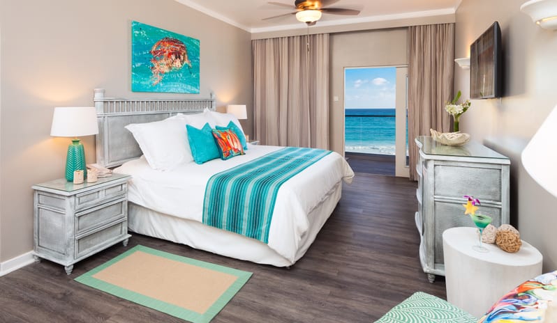 Classic Ocean View Coastal-inspired resort bedroom interior displaying whitewashed wooden furniture, teal accents, a ceiling fan, and direct views of blue ocean waters at Sea Breeze Beach House by Kenwood Travel