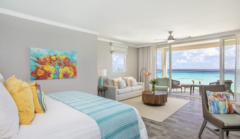 Luxury Junior Suite Ocean Front Coastal resort bedroom suite showing vibrant floral canvas artwork, soft neutral sofa seating, and direct views of turquoise ocean waters through large balcony doors at Sea Breeze Beach House by Kenwood Travel
