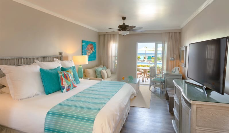 Classic Garden View Tropical hotel bedroom interior decorated in teal and beige tones, complete with a cozy lounge seating area, desk, and view of an outdoor pool deck at Sea Breeze Beach House by Kenwood Travel