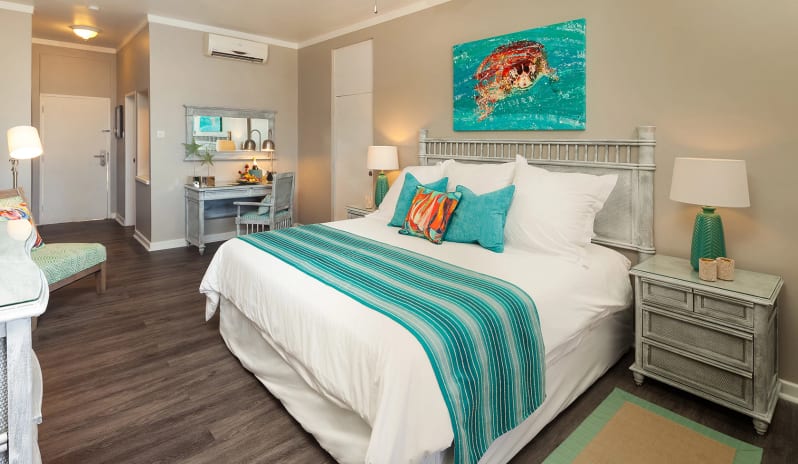 Classic Garden View Tropical resort suite interior displaying a white king bed, turquoise throw pillows, a rustic vanity desk, and coastal wall art at Sea Breeze Beach house by Kenwood Travel