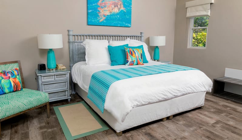 Classic Suite Two Bedroom Coastal resort king suite interior with whitewashed wooden furniture, bright turquoise throw pillows, dark wood-plank flooring, and a window with garden views at Sea Breeze Beach House by Kenwood Travel