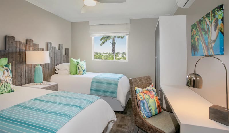 Luxury Suite Two Bedroom Ocean View Modern twin bedroom interior in a tropical suite showcasing colorful wildlife wall art, a woven chair with a patterned cushion, and a window with palm tree views at Sea Breeze Beach House by Kenwood Travel