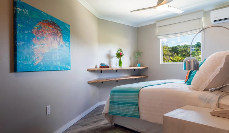 Luxury Suite Two Bedroom Ocean Front Coastal resort bedroom interior with light beige walls, a white nightstand, chrome reading lamp, modern ceiling fan, and vibrant turquoise wall decor  at Sea Breeze Beach House by Kenwood Travel