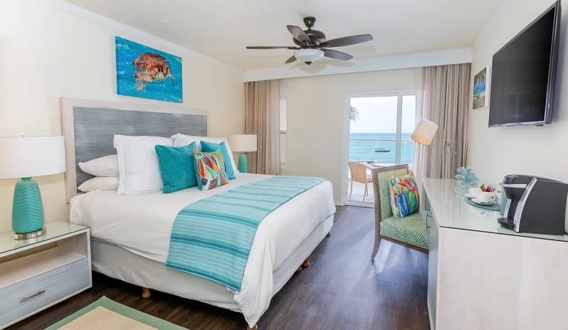 Classic Ocean Front Tropical beachfront resort suite displaying a king-size bed, teal bedside lamps, a ceiling fan, and direct views of the blue sea through patio doors at Sea Breeze Beach House by Kenwood Travel