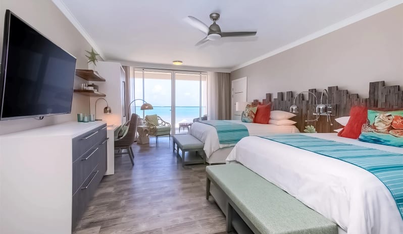 Luxury Suite Two Bedroom Ocean Front Luxury resort double bedroom with wood-look flooring, modern white console, large wall-mounted flat-screen TV, desk workstation, ceiling fan, and direct views of the ocean at Sea Breeze Beach House by Kenwood Travel
