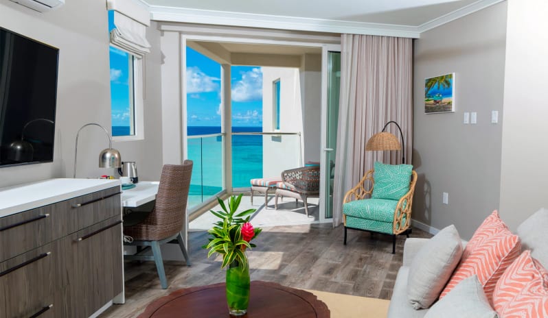 Luxury Suite Three Bedroom Ocean Front Spacious beachfront suite lounge with light grey walls, a plush couch with coral cushions, a solid wood coffee table with fresh tropical flowers, wood-style flooring, and floor-to-ceiling glass doors leading out to a scenic ocean-view balcony at Sea Breeze Beach House by Kenwood Travel