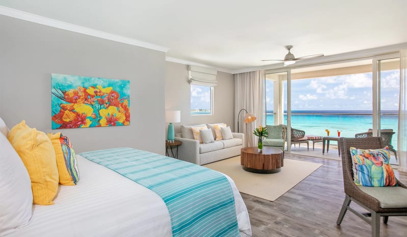 Luxury Suite Two Bedroom Ocean Front Coastal resort bedroom interior showing vibrant floral wall art, a soft white couch, a round wooden coffee table, and panoramic ocean views through floor-to-ceiling balcony doors at Sea Breeze Beach House by Kenwood Travel