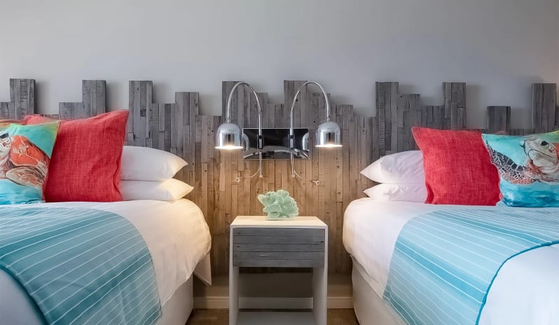 Luxury Suite Two Bedroom Ocean Front Symmetrical view of two plush hotel beds with white linens, light blue striped runners, sea turtle accent pillows, and dual chrome reading lights fixed to a rustic wood panel feature wall at Sea Breeze Beach House by Kenwood Travel