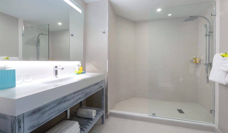 Luxury Suite Two Bedroom Ocean Front Coastal hotel bathroom interior displaying a white floating sink vanity, plush white towels, bright backlit mirror, and a seamless tiled walk-in shower at Sea Breeze Beach House by Kenwood Travel