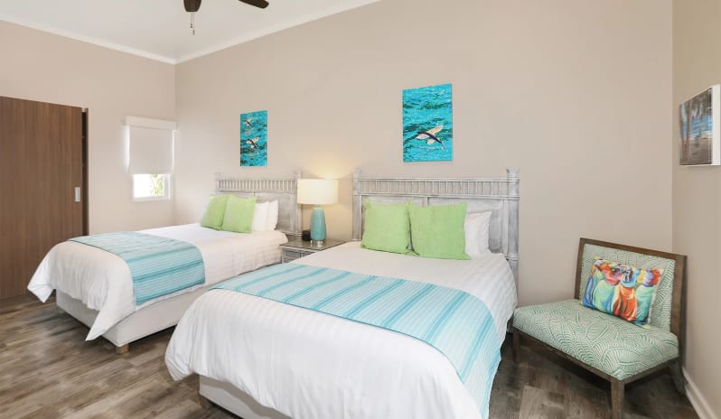 Classic Suite Two Bedroom Tropical hotel double bedroom interior with light wood-style flooring, whitewashed headboards, colorful aquatic art pieces, and a comfortable corner armchair at Sea Breeze Beach House by Kenwood Travel