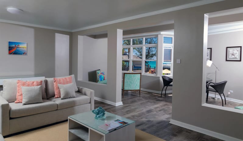 Classic Suite Two Bedroom Modern coastal suite living area with neutral grey walls, wood-style flooring, a beige couch, and large windows looking out toward a tropical resort pool and ocean at Sea Breeze Beach House by Kenwood Travel