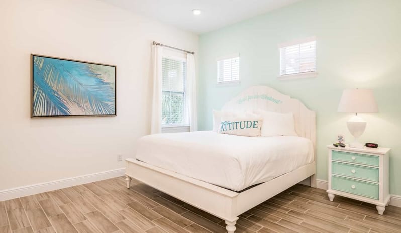 Three Bedroom Villa Bedroom with a white bed frame, turquoise-accented nightstand, table lamp, and blue palm artwork at Margaritaville Resort Orlando by Kenwood Travel