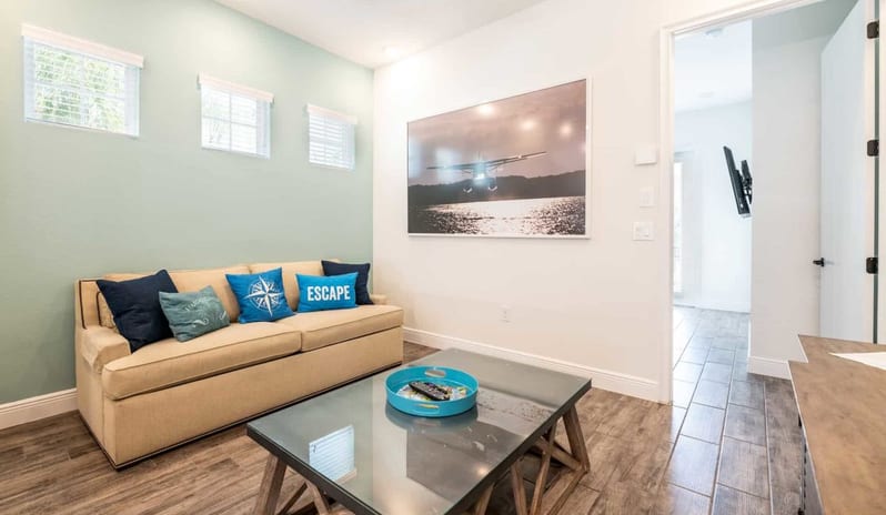 Three Bedroom Villa Small sitting room featuring a beige couch with blue pillows, a glass-top coffee table, and large airplane artwork at Margaritaville Resort Orlando by Kenwood Travel