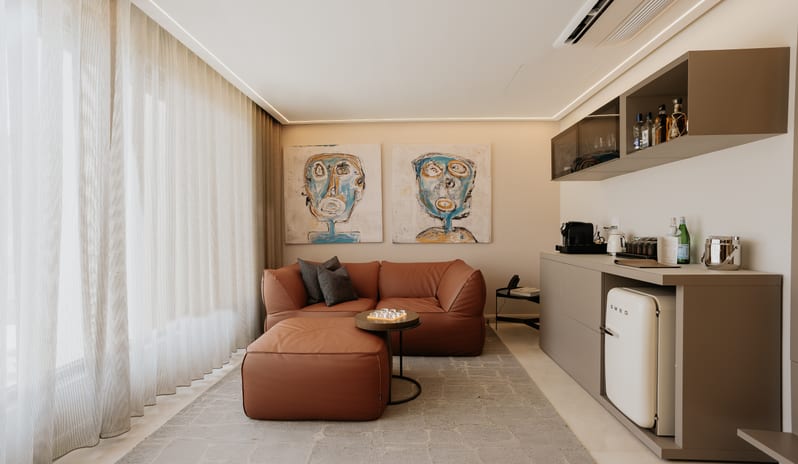 Blue Monkey Signature One Bedroom Suite  Luxury hotel suite living area featuring a brown sectional sofa, ottoman, artwork, shear curtains, and a fully stocked minibar setup at Blue Monkey Hotel & Beach Club by Kenwood Travel