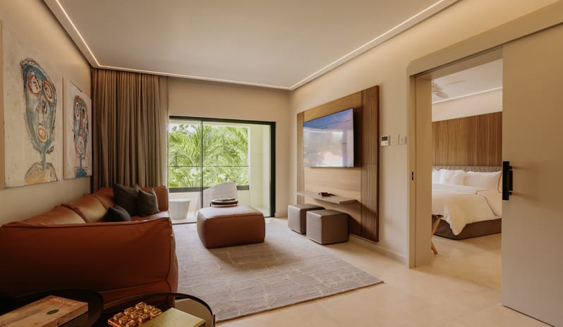 Luxury Pool Suite Spacious luxury hotel suite living room with a leather couch, wall-mounted TV, ottoman, and sliding door leading into an adjoining bedroom at Blue Monkey Hotel & Beach Club by Kenwood Travel
