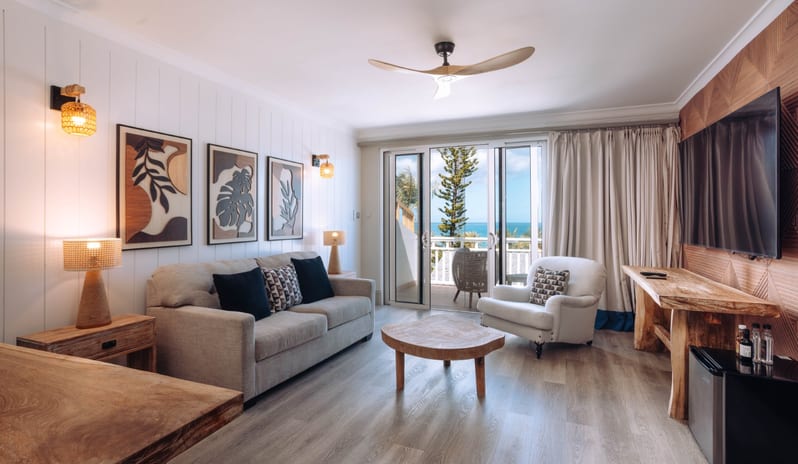 Hillside Luxury Modern coastal living room featuring a beige sofa, wooden coffee table, wall art, and glass sliding doors opening to an ocean view balcony at Blue Water Resort & Spa by Kenwood Travel