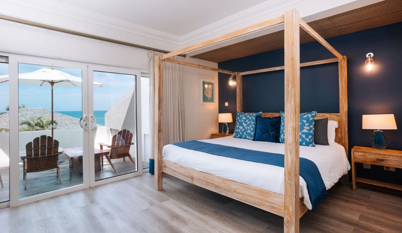 Hillside Luxury Modern coastal bedroom featuring a light wood four-poster canopy bed, deep navy blue accent wall, and sliding glass doors opening to an ocean-view balcony at Blue Waters Resort & Spa by Kenwood Travel