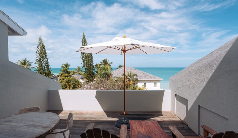 Hillside Luxury Room Spacious outdoor sun deck featuring wooden patio furniture, a white patio umbrella, and scenic views of the turquoise ocean and pine trees at Blue Waters Resort & Spa by Kenwood Travel