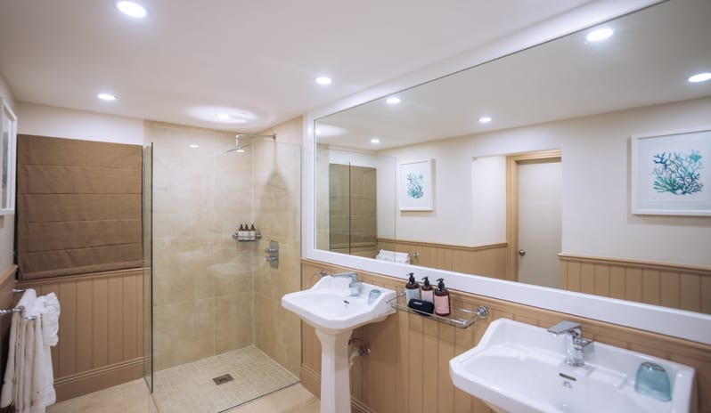 Hillside Luxury Room Spacious coastal bathroom featuring double pedestal sinks, a large framed mirror, beige wall wainscoting, and a walk-in glass rain shower at Blue Waters Resort & Spa by Kenwood Travel