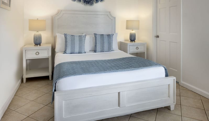 Prime minister Manor Coastal style resort bedroom with light wood queen bed frame, blue runner blanket, dual bedside tables, and soft warm lighting at Windjammers Landing by Kenwood travel