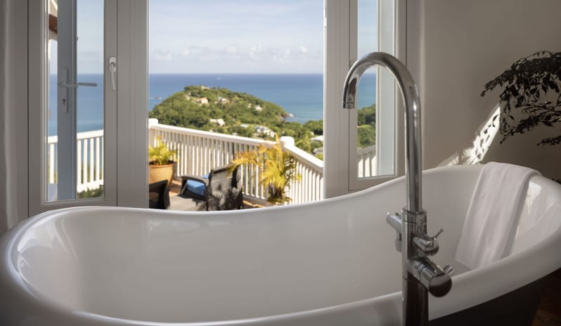Dukes Manor White freestanding soaking tub with a gooseneck chrome faucet positioned next to open balcony doors framing a panoramic ocean and coastal hill view at Windjammers Landing by Kenwood travel