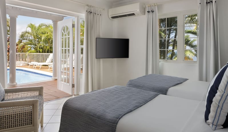 Prime minister Manor Bright villa guest bedroom with twin beds, blue woven throws, a wall-mounted TV, and open french doors leading to a poolside patio at Windjammers Landing by Kenwood travel