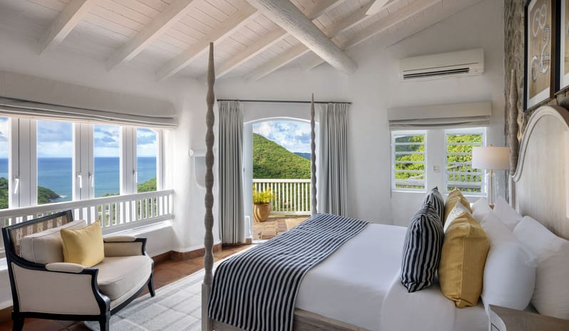 Dukes Manor Spacious luxury villa bedroom with vaulted white wood ceilings, a four-poster bed, cozy armchair with a yellow pillow, and large windows looking out at the ocean and green hills at Windjammers Landing by Kenwood travel