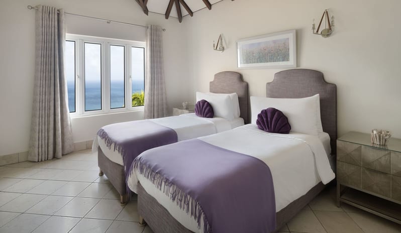 Royal Manor Spacious Cozy guest bedroom featuring two twin beds with upholstered grey headboards, soft purple fringed blankets, fan-shaped decorative pillows, and large windows looking out to the ocean at Windjammers Landing by Kenwood travel
