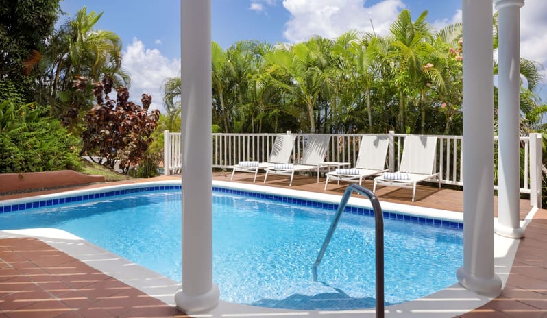 Prime minister Manor Sunlit resort swimming pool surrounded by tiled patio columns, white lounge chairs on a wooden deck, and lush tropical palm trees at Windjammers Landing by Kenwood travel