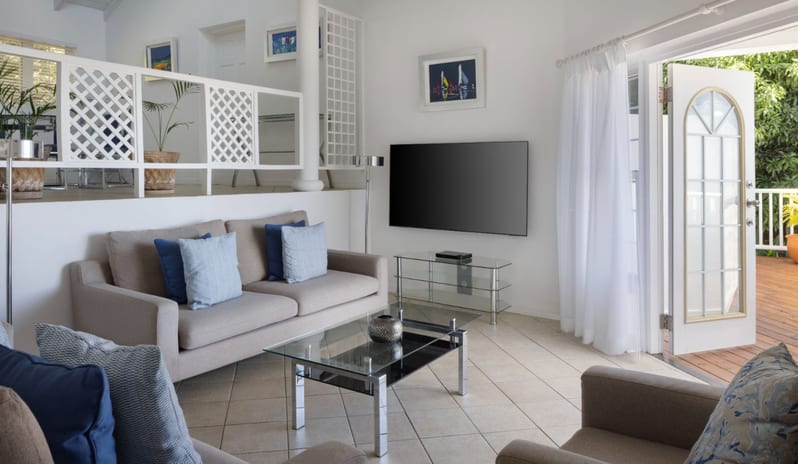 Prime minister Manor Luxury beach villa living room with modern seating, wall-mounted flat-screen TV, white lattice balcony railing, and sunlit outdoor terrace access at Windjammers Landing by Kenwood travel