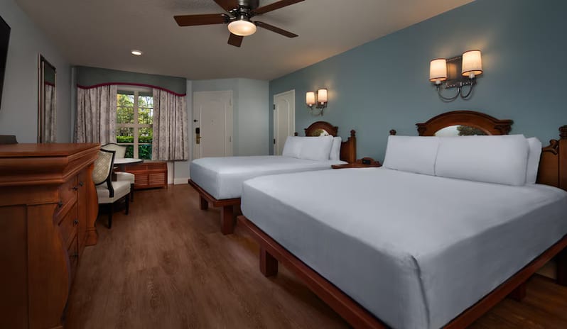 Standard Location Room with 5th Sleeper - Spacious hotel room featuring two queen beds with wooden frames, light blue walls, soft wall sconces, and a ceiling fan at Disney's Port Orleans Resort - Riverside by  Kenwood travel
