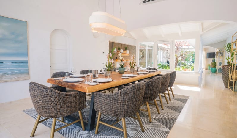 Presidential Manor Spacious luxury villa dining area with a long live-edge wooden table, dark woven armchairs, modern pendant light, geometric rug, and sea artwork at Windjammers Landing by Kenwood travel