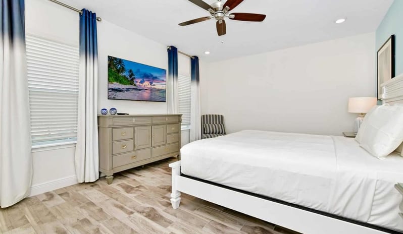 Four Bedroom Villa Master bedroom with a white king bed, dresser, wall-mounted TV, and blue ombre drapes at Margaritaville Resort Orlando by Kenwood Travel