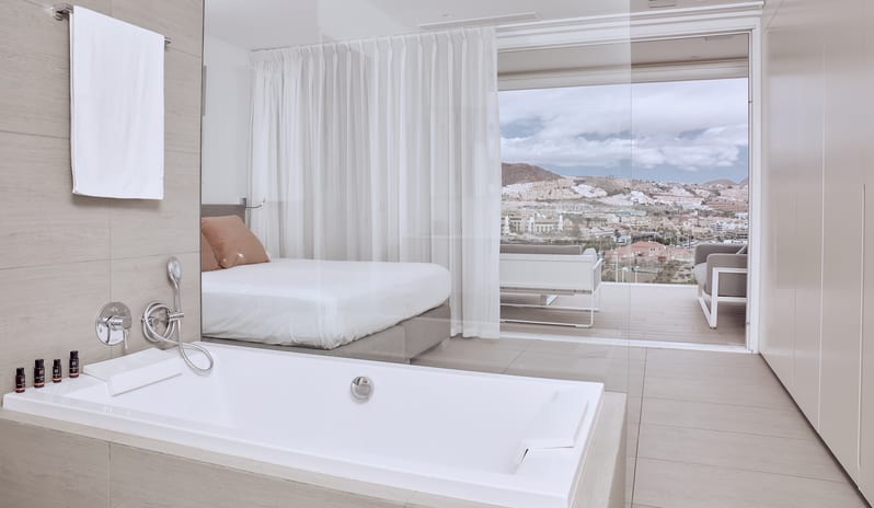 Serenity Allegra Spacious resort suite bathtub next to a glass partition showing a bedroom and a balcony view of a hillside town at Domes Baobab by Kenwood Travel