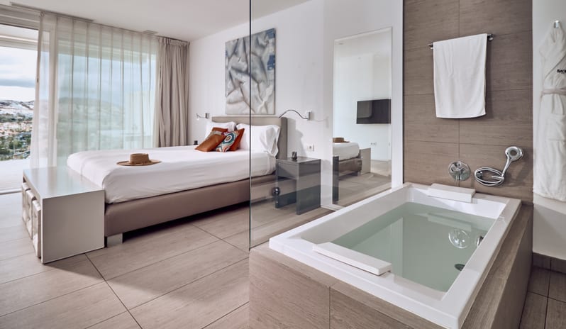 Serenity Essence Luxurious hotel suite featuring a king-size bed with straw hat decor, plush white bathrobe, and a deep spa bathtub overlooking the room at Domes Baobab by Kenwood Travel