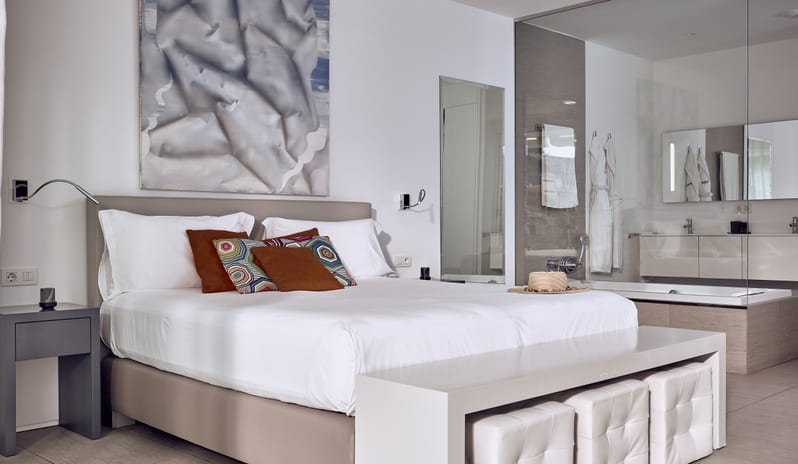 Serenity Essence Luxury resort suite featuring a plush king bed, sleek dark nightstand, and a seamless glass-wall view into a spa bathroom with plush bathrobes and a bathtub at Domes Baobab by Kenwood Travel
