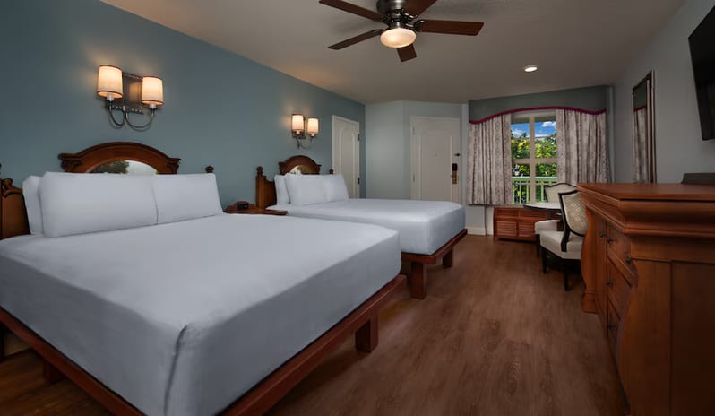 Preferred Location Room - Wide interior view of a luxury hotel room featuring two queen-size beds with crisp white linens, dark wooden furniture, soft blue walls, wall-mounted lamps, and a window with outdoor views at Disney's Port Orleans Resort - Riverside by  Kenwood travel