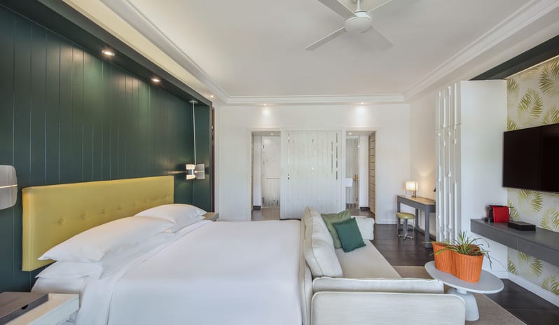 Junior Suite living area king bed with yellow headboard, beige sofa, vanity desk, ceiling fan, and green accent panel wall at Long Beach Mauritius by Kenwood Travel