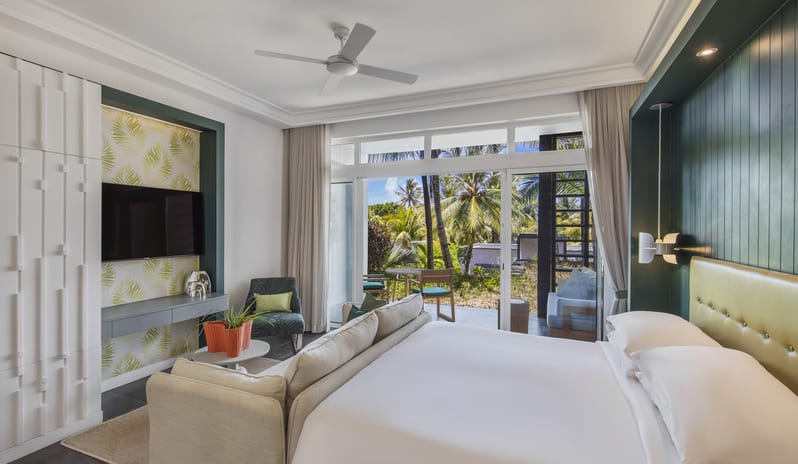 Junior Suite living area king bed, sofa lounge, mounted TV, and sliding glass doors opening to a private garden terrace surrounded by palm trees at Long Beach Mauritius by Kenwood Travel