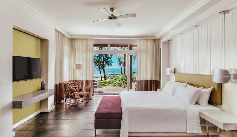 Long Beach Executive Suite king-size bed, upholstered bench, wall-mounted TV console, and wide glass doors opening to an ocean-view terrace at Long Beach Mauritius by Kenwood Travel