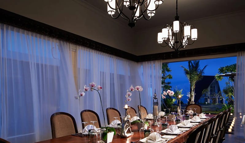 Royal Samabe Residence dining table set for dinner with chandeliers, orchid centerpieces, and sheer curtain windows at dusk at Samabe Bali Suites and Villas by Kenwood Travel