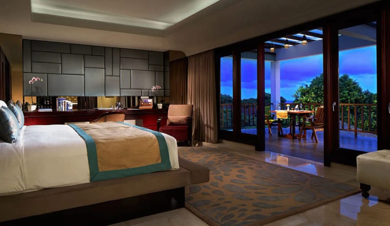 Royal Samabe Residence master bedroom with king bed and open glass doors leading to a private balcony with outdoor dining at dusk at Samabe Bali Suites and Villas by Kenwood Travel