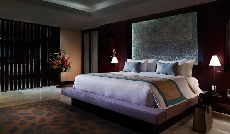 Royal Samabe Residence plush king-size bed, illuminated textured headboard panel, bedside lamps, and decorative floral arrangement at Samabe Bali Suites and Villas by Kenwood Travel