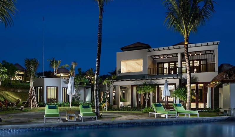 Royal Samabe Residence Illuminated luxury resort villa at night featuring poolside sun loungers, palm trees, and swimming pool against a deep blue sky at Samabe Bali Suites and Villas by Kenwood Travel