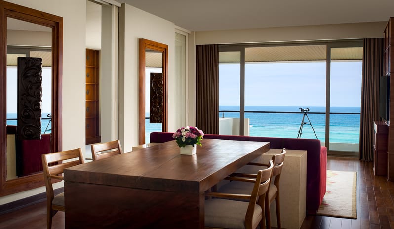 Ocean Front Family Suite dining room featuring a solid wood dining table with floral centerpiece and panoramic ocean views through floor-to-ceiling glass doors at Samabe Suites & Villas by Kenwood Travel
