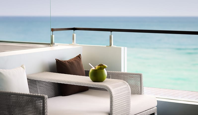 Ocean Front Family Suite private balcony featuring a woven wicker daybed with fresh drinking coconut and panoramic ocean views at Samabe Suites & Villas by Kenwood Travel